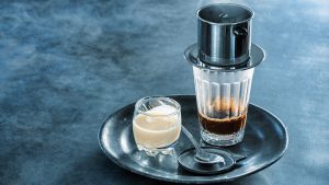 capture of coffee being prepared in the classic Vietnamese style. The still steaming metal drip filter, known as phin cà phê sits on top of a glass, which sits on a small tray alongside a smaller glass containing sweetened condensed milk. This simple setup is the beginning for classic Vietnamese iced coffee, as well as several other coffee drinks.