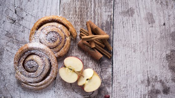 Two cinnamon buns topped with icing sugar and decorated with apple slices and cinnamon sticks