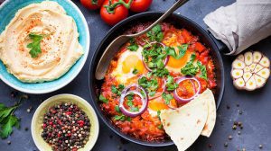 schnelle Gerichte, Shakshuka with pita bread in pan, hummus in bowl on rustic background. Middle eastern traditional dishes. Fried eggs with vegetables. Top view. Space for text. Middle eastern style breakfast or lunch