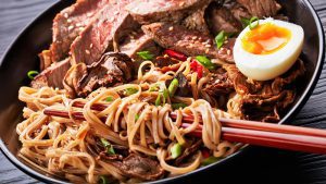 a bowl of soba noodles with sliced roast beef steak, shiitake mushrooms, half of hard boiled egg and fried vegetables with chopsticks, asian cuisine, view from above, close-up