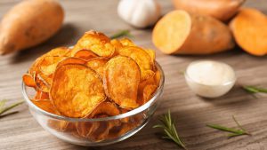 Delicious sweet potato chips in bowl, rosemary and sauce on table. Space for text