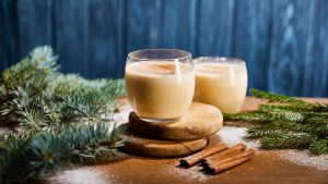 delicious eggnog cocktail on round wooden boards near spruce branches, cinnamon sticks and scattered sugar powder on blue textured background