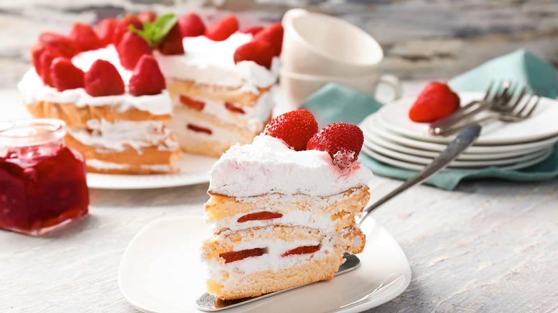 Stück Erdbeertorte auf einem Teller, im Hintergrund gedeckte Kaffeetafel