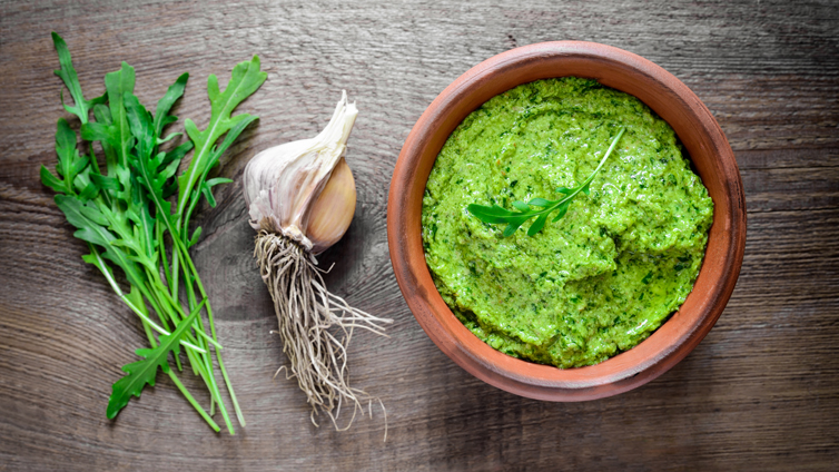 Rucola-Pesto in kleinem Tontopf neben Bund Rucola und Knoblauch