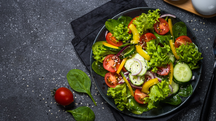Ein grüner Blattsalat mit Gurke, Paprika, Tomaten ist auf einem dunklen, tiefen Teller angerichtet.