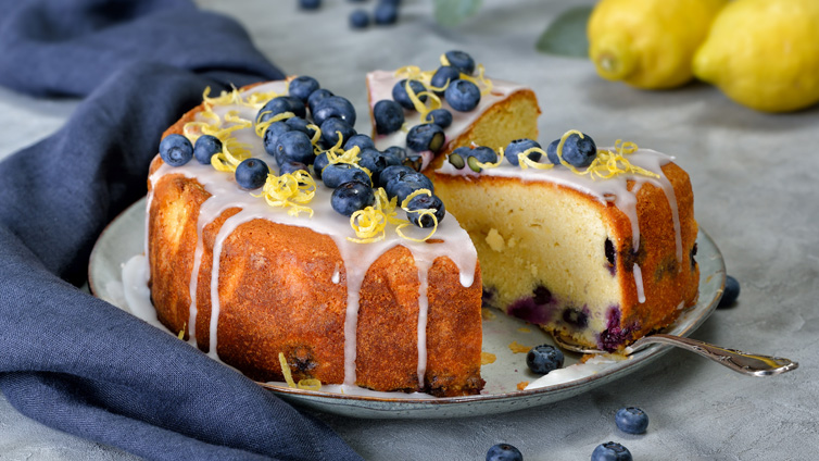 Heidelbeerkuchen mit Zuckerguss, Heidelbeeren und Zitronenzeste steht angeschnitten auf einem großen Teller.