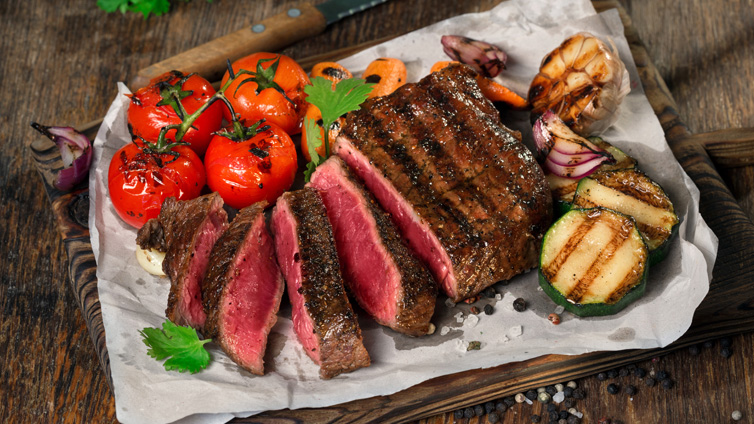 Ein gegrilltes und angeschnittenes Stück Roastbeef liegt mit Grillgemüse auf einem Pergamentpapier auf einem Holzbrett.