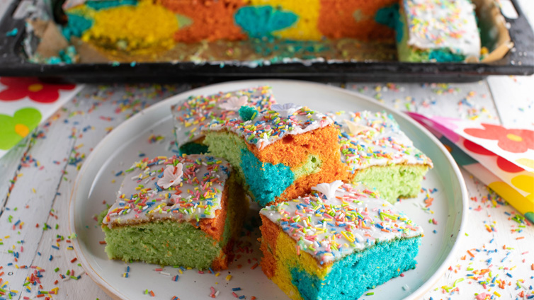 Mehrere Stücke bunter Papageienkuchen mit Streuseln liegen auf einem weißen Teller übereinandergestapelt, im Hintergrund steht ein Kuchenblech.