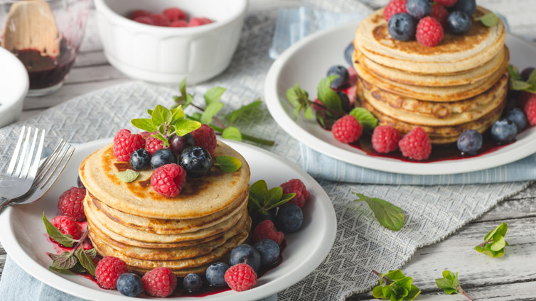 Zwei Teller mit je einem Stapel Pancakes, Beeren und Minze stehen auf einem Tisch.