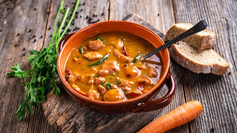 Ein Löffel steckt in einer Gyrossuppe, die in einer Schüssel auf einem Holzbrett serviert wird.