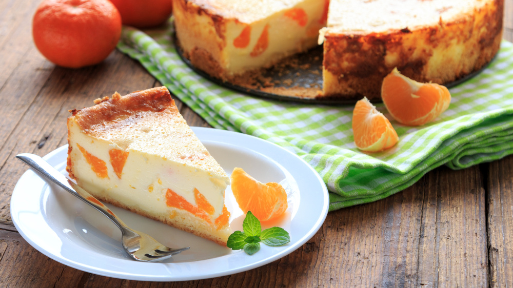 Ein Stück Käsekuchen mit Mandarinen liegt auf einem Teller neben Mandarinen.